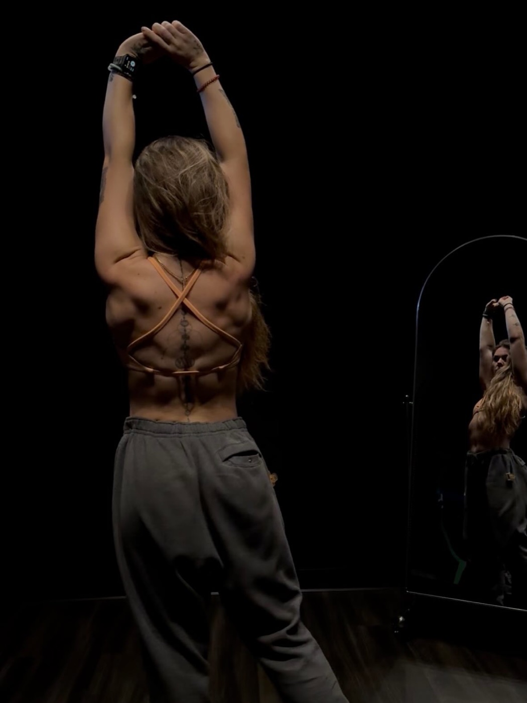 Kira Marlow stretching overhead in a dark studio, seen from behind